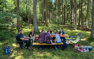 Neun Personen sitzen an und um eine Biertischgarnitur im Wald und essen