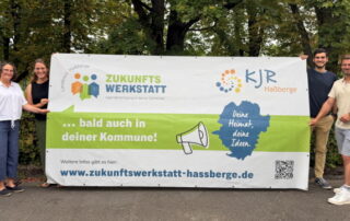 Susanne Makowski, Eva Pfeil, Peter Weisheit und Thomas Wagenhäuser stehen vor dem Park der Promenade neben dem Bauzaunbanner der Zukunftswerkstätten und werben für Helferinnen und Helfer 2026